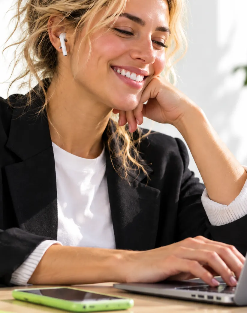A smiling woman in a blazer and earbuds typing on her laptop at a sunny desk
